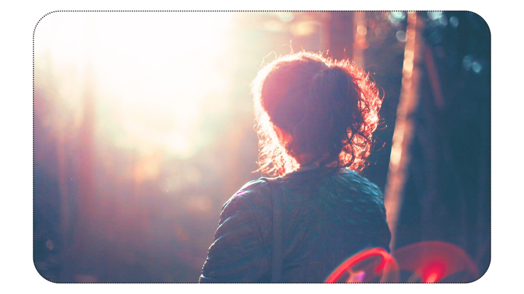 Woman, facing away, looking into the sunshine peaking through the woods. 